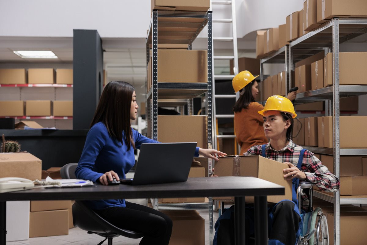 warehouse-manager-coordinating-logistics-operators-team-from-desk-checking-parcel-packing-quality-asian-storehouse-worker-wheelchair-giving-cardboard-box-coworker-table-1-.jpg